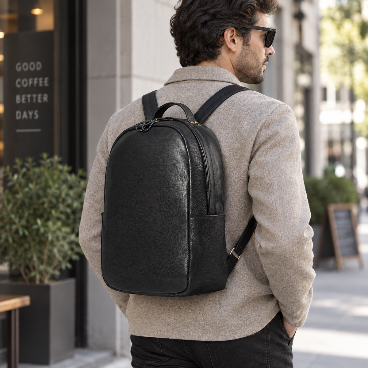 A man wearing a black leather backpack, seen from behind as he walks down a city street. He is wearing a beige sweater and black pants, with a coffee shop storefront in the background.