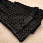 Black woven leather accessory with a strap and snap closure, photographed on a soft beige textured surface.