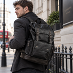 A man wearing a dark gray coat and black jeans carries a large black leather backpack with brown leather trim and brass-tone hardware, photographed on a city street with buildings, a black railing, and a red double-decker bus in the background.