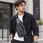 A man wearing a black crossbody sling bag with multiple zippered pockets and a gold-tone chain detail, styled over a white t-shirt and black overshirt in an urban setting.