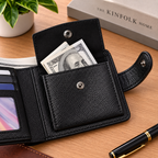 A black textured leather bi-fold wallet shown open on a wooden desk, with cash and cards visible inside. A pen, a small potted plant, and a book are placed around it as styled props.