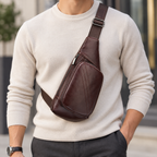A man wearing a cream-colored knit sweater and dark gray trousers, carrying a dark brown leather sling bag across his chest. The bag features a wide adjustable strap and a front zippered pocket.