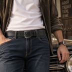A close-up lifestyle shot of a man wearing a black leather belt with a silver-toned buckle, styled with a white t-shirt, brown overshirt, and dark blue jeans, leaning against a vintage car.