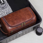 Brown leather toiletry bag on top of an open suitcase with a white surface underneath.